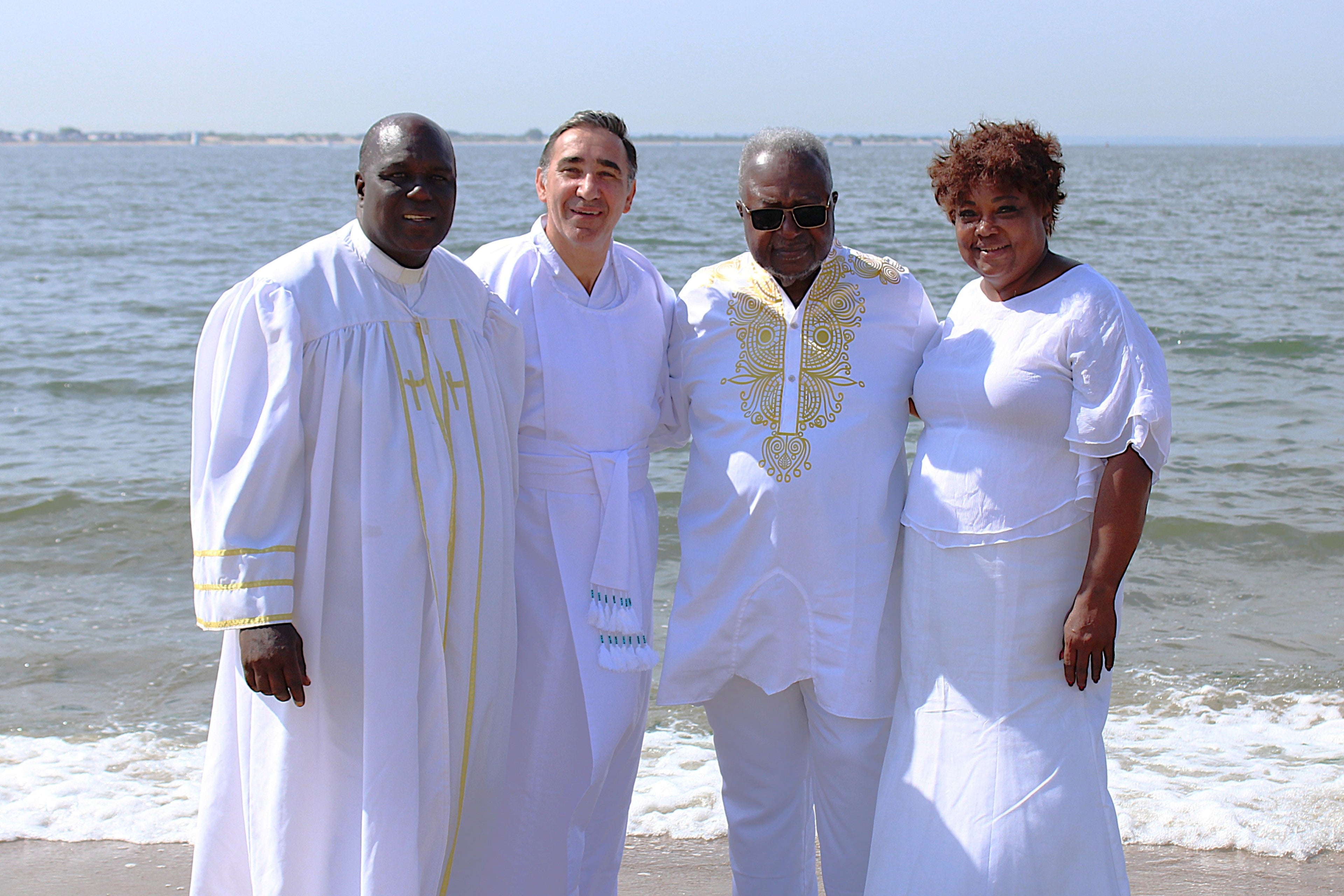 Four people in white outfits standing on a beach with ocean in the background